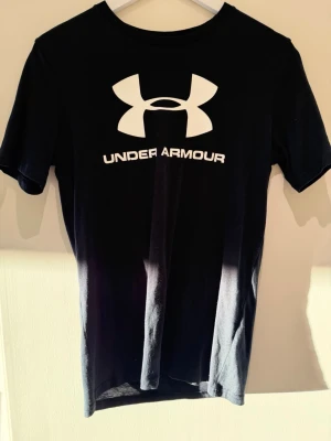 Svart Under Armour t-shirt XS - Säljer en svart t-shirt från Under Armour i storlek XS. T-shirten har korta ärmar, rund halsringning och stor vit logga samt text på bröstet. Modellen är loose fit och tillverkad i ett mjukt bomullsmaterial som passar perfekt till träning.
