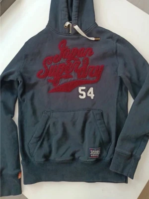Blå Superdry hoodie med röd logga - Mörkblå hoodie från Superdry med stor röd broderad 'Japan Superdry' logga och siffran 54 på bröstet. Klassisk känguruficka framtill, huva med vita dragsnören och patch vid fickan. Perfekt för en avslappnad streetwear-look.