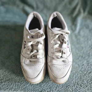 Vita Fila sneakers med logga - Säljer ett par klassiska vita sneakers från Fila med tydlig logga i blått och rött på sidan. Skorna har en chunky sula, lågt skaft och är tillverkade i syntetmaterial och textil. Perfekta för en sportig och avslappnad stil.