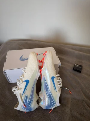 Nike vita fotbollsskor med blå swoosh - Säljer ett par vita Nike fotbollsskor med blå swoosh och detaljer i neonorange på dobbarna. Skorna har en modern, strömlinjeformad design och är tillverkade i syntetmaterial för bästa grepp och känsla på planen. Perfekta för dig som vill sticka ut på matchen!