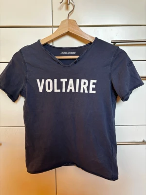 Blå Zadig & Voltaire t-shirt storlek 138/ XXXXS - Mörkblå t-shirt från Zadig & Voltaire med vit text 'VOLTAIRE' framtill. T-shirten har korta ärmar, rund halsringning med råa kanter och små knappar vid halsen. Tillverkad i mjuk bomull för en avslappnad look. Storlek 138/ 10 år/ XXXXS