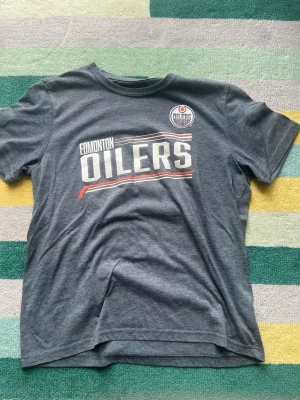 Edmonton Oilers t-shirt McDavid - Snygg mörkgrå Edmonton Oilers t-shirt med tryck fram och stort nummer 97 samt McDavid på ryggen. Klassisk rund hals och korta ärmar. Perfekt för dig som gillar hockey och vill visa ditt stöd för laget.