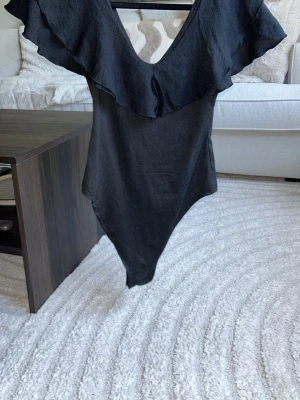 Svart body med volangdetalj - Snygg svart body med djup v-ringning och bred volang runt halsringningen. Kroppsnära passform och mjukt material som känns bekvämt mot huden. Perfekt att styla med jeans eller kjol för en trendig look.