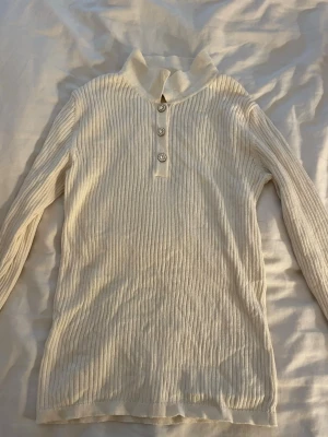 Vit ribbstickad polotröja - Snygg vit ribbstickad polotröja med långa ärmar och tre dekorativa knappar framtill. Tröjan har en hög krage och är gjord i ett mjukt, stretchigt material som sitter skönt på kroppen. Perfekt för lager på lager och enkel att matcha.