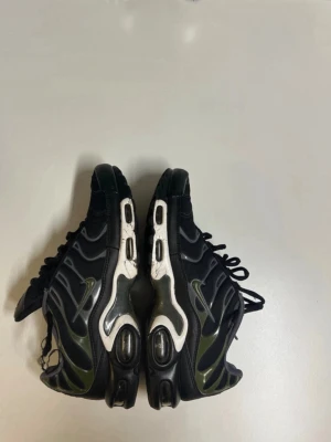 Nike Air Max Plus svart/grön - Nike Air Max Plus sneakers i svart och olivgrönt med vita detaljer. Skorna har den ikoniska vågiga designen, synlig Air-dämpning i sulan och svarta snören. Materialet är en mix av mesh och syntet. Perfekta för dig som gillar streetwear och sportig stil.
