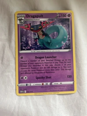 Dragapult Pokémonkort - Dragapult Pokémonkort med lila bakgrund och gul ram. Kortet har illustration av Dragapult, en drake med turkos kropp och röda detaljer. Kortet har 150 HP och attacker som Dragon Launcher och Spooky Shot. Perfekt för samlare eller Pokémon-fans.