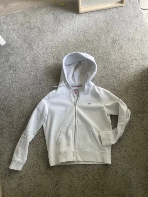 Vit hoodie från Tommy Jeans - Snygg vit hoodie från Tommy Jeans med dragkedja och klassisk logga på bröstet. Hoodien har huva med dragsko och en stor magficka framtill. Perfekt för en clean och stilren look. Tillverkad i mjukt bomullsmaterial.