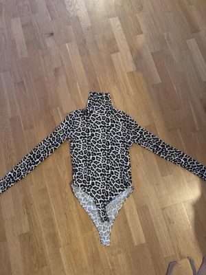 Leopardmönstrad body med polokrage - Snygg body med leopardmönster och lång ärm. Hög polokrage och figurnära passform, perfekt för att skapa en trendig look. Materialet är stretchigt och bekvämt, vilket gör den enkel att bära hela dagen. Passar dig som gillar djurmönster och vill sticka ut.