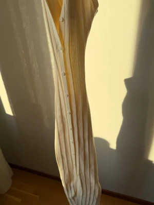 Lång ribbad klänning i ljusbeige - Lång ärmlös ribbad klänning i ljusbeige med knappar framtill. Klänningen har en smickrande slim passform och är tillverkad i ett mjukt, stretchigt material som ger en bekväm känsla. Perfekt för varmare dagar och enkel att styla med accessoarer.OBS den har en fin öppen rygg