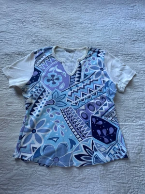 Mönstrad blå och vit t-shirt - Unik t-shirt med korta vita ärmar och ett färgstarkt blått och lila mönster framtill och baktill. T-shirten har v-ringning och är tillverkad i mjuk bomull, perfekt för dig som gillar retrovibbar och grafiska tryck. Bröst 96cm, bomull 100%