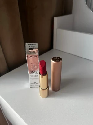 Lancôme rött läppstift 888 Kind of Sexy - Snyggt rött läppstift från Lancôme i nyansen 888 Kind of Sexy. Kommer i en lyxig guldig hylsa med brunt lock och vacker rosdetalj på toppen. Förpackningen är elegant och stilren, perfekt för dig som vill ha en klassisk röd färg på läpparna.