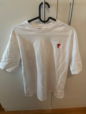 Vit t-shirt med rött hjärta Ami - Klassisk vit t-shirt från AMI med ett litet rött hjärta och A broderat på bröstet. T-shirten har rund hals, normal passform och korta ärmar. Tillverkad i mjuk bomull som känns skön mot huden. Perfekt till jeans eller shorts för en clean look. Lite mer oversized