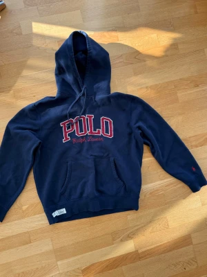 Mörkblå hoodie från Polo Ralph Lauren - Mörkblå hoodie från Polo Ralph Lauren med stor broderad POLO-text i rött och vitt på bröstet. Klassisk känguruficka framtill, dragsko i huvan och liten röd logga på ärmen. 