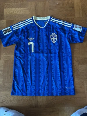 Blå retro fotbollströja Adidas - Säljer en blå retro fotbollströja från Adidas med nummer 7 på bröstet och klassiskt mönster. Tröjan har vita detaljer, broderad svensk landslagssköld och Adidas-logga. Korta ärmar med vita ränder på axlarna och V-ringad krage.
