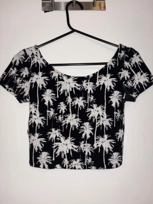 Svart croppad topp med palmtryck H&M - Snygg croppad topp från H&M Divided i svart med vitt palmtryck. Toppen har rund halsringning och korta ärmar, perfekt för en avslappnad och trendig look. Tillverkad i mjuk bomullsmix som känns skön mot huden.