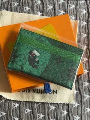 Grön plånbok med golfformat  - Snygg grön korthållare från L med svart monogrammönster och coolt motiv framtill. Tillverkad i tåligt läder och har plats för flera kort. Perfekt accessoar för dig som gillar stilrena och unika detaljer. Plånboken skickas med det på bilderna det är en golf edition på plånboken.
