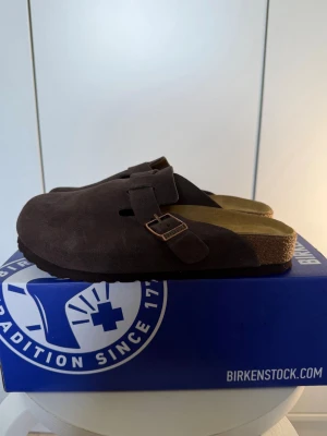 Birkenstock Boston BS mocka tofflor - Mörkbruna Birkenstock Boston BS tofflor i mocka med justerbart spänne och klassisk korksula. Skön fotbädd och stilren design som passar perfekt för dig som gillar en avslappnad look. Färg: mocha.