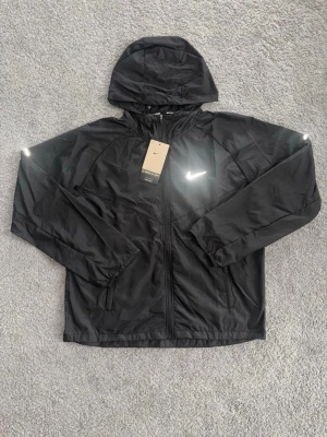 Svart Nike Miller vindjacka  - Snygg svart vindjacka från Nike med reflekterande logga på bröstet och detaljer på axlarna. Jackan har huva, dragkedja framtill och två sidofickor. Tillverkad i lätt och tåligt material, perfekt för löpning eller promenader. Nike Running-tryck på insidan.