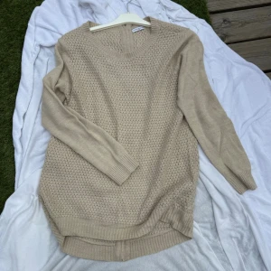 Beige stickad tröja Jacqueline de Yong - Beige stickad tröja från Jacqueline de Yong med rund halsringning och lång ärm. Tröjan har ett diskret stickat mönster framtill och ribbade muddar vid ärmslut och nederkant. Perfekt för lager-på-lager och enkel att matcha.