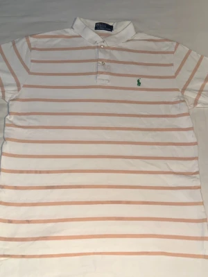 Randig pikétröja från Polo Ralph Lauren - Klassisk vit pikétröja från Polo Ralph Lauren med ljusrosa horisontella ränder och grönt broderat logomärke på bröstet. Tröjan har krage, knäppning med två knappar och korta ärmar. Perfekt för en avslappnad och stilren look. Står som storlek M men är som en mindre L också. Fråga om mått vid behov :) 