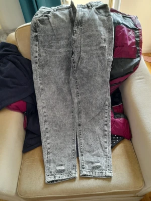 Grå acid wash mom jeans - Säljer ett par grå acid wash mom jeans med hög midja och raka ben. Jeansen har slitningar vid bensluten och klassisk femficksdesign. Perfekta för en avslappnad och trendig look.