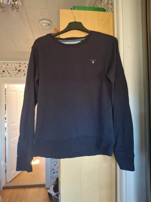 Mörkblå sweatshirt från Gant - En stilren mörkblå sweatshirt från Gant med rund halsringning och diskret logga på bröstet. Tröjan har långa ärmar och ribbade muddar vid ärmslut och nederkant. Perfekt för en avslappnad och snygg look. Använd få gånger så är I ett väldigt bra skick!! Passar storlek S/M