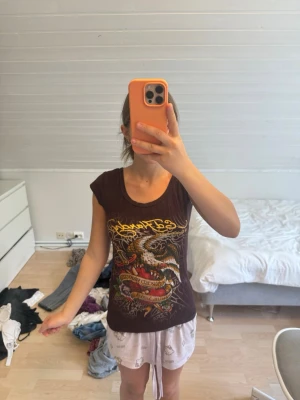 Brun t-shirt med draktryck och slitsad rygg - Cool brun t-shirt med färgglatt drakmotiv och text framtill. T-shirten har korta ärmar, rund halsringning och unik slitsad detalj i ryggen. Perfekt för dig som gillar edgy streetstyle och vill sticka ut. Materialet känns mjukt och skönt mot huden.