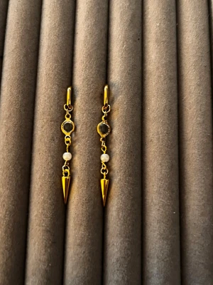 Guldpläterade örhängen med pärlor och spikes - Säljer ett par trendiga guldpläterade örhängen med små vita pärlor och svarta stenar. Örhängena har en cool hängande design med spetsiga spikes längst ner. Perfekta för dig som vill ha något unikt och edgy till din stil.