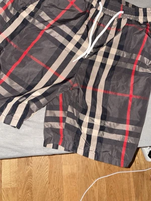 Rutig shorts med snörning - Snygga rutiga shorts med svart, beige och röda detaljer samt vit snörning i midjan. Perfekta för sommaren och har en avslappnad passform. Klassisk design som sticker ut med sitt mönster.