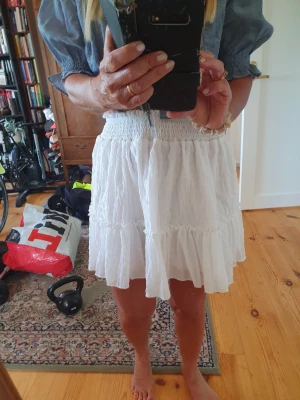 Vit volangkjol med smock - Säljer en söt vit kortkjol med bred smock i midjan och volangdetaljer. Kjolen har en luftig och lätt känsla, perfekt för varma dagar. Passar till både sneakers och sandaler för en chill look.
