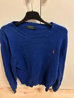 Blå stickad tröja Polo Ralph Lauren - Klassisk blå stickad tröja från Polo Ralph Lauren med rund halsringning och ribbade muddar. Tröjan har den ikoniska röda logotypen broderad på bröstet. Perfekt för lager-på-lager och enkel att matcha med jeans.