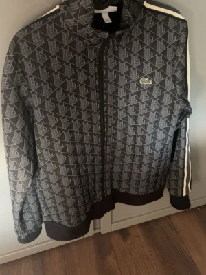 Mönstrad zip-hoodie från Lacoste - Säljer en svart och grå zip-hoodie från Lacoste med ett grafiskt mönster och den klassiska krokodilloggan på bröstet. Tröjan har vita ränder längs ärmarna och muddar vid ärmslut och nederkant. Perfekt för dig som gillar streetwear och snygga detaljer.