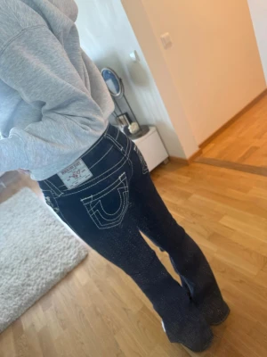 Blå True Religion jeans med utsvängda ben - Säljer ett par mörkblå jeans från True Religion med ikoniska kontrastsömmar och utsvängda ben. Jeansen har låg midja och stora bakfickor med tydlig logga. Perfekta för dig som gillar en retro och trendig look.