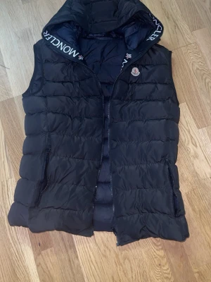 Svart dunväst från Moncler - Snygg svart dunväst  med huva och dragkedja framtill. Västen har ogga på bröstet och märkning på huvkanten. Perfekt för lager-på-lager och håller dig varm under kyliga dagar.