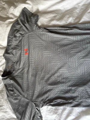 Grå tränings-tshirt från Under Armour - Snygg grå tränings-tshirt från Under Armour med diskret geometriskt mönster och orange logga på bröstet. Tillverkad i ett lätt och snabbtorkande funktionsmaterial som passar perfekt till gymmet eller löprundan. Rund hals och korta ärmar för maximal rörelsefrihet.