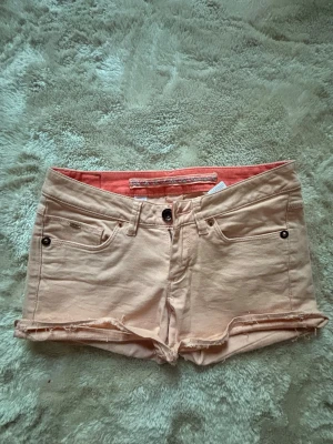 Rosa shorts med - Säljer ett par beiga shorts med rå kant och klassisk femficksdesign. Shortsen har knapp och dragkedja framtill samt bälteshällor. Insidan har en snygg korallfärgad detalj och ett flätat band i midjan. Perfekta för varma dagar!