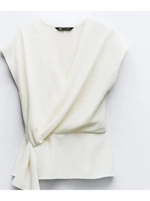 Zara omlott topp - Stilren vit/creme omlottblus från Zara med knyt i sidan och v-ringad hals. Blusen är ärmlös och har en mjuk, flowy passform som ger en avslappnad men chic look.  Använd fåtal gånger.