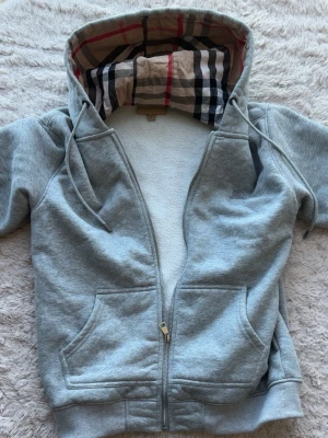 Grå zip-hoodie från Burberry - Snygg grå hoodie med dragkedja från Burberry. Klassisk rutmönstrad huva och broderad logga på bröstet. Mjuk bomullsblandning, två fickor fram och dragsko i huvan. Perfekt för en chill och stilren look.