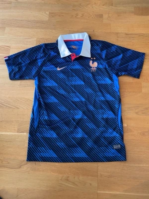 Blå Nike Frankrike fotbollströja - Snygg blå fotbollströja från Nike med Frankrikes landslagsmärke och två stjärnor på bröstet. Tröjan har ett diagonalrandigt mönster i olika blå nyanser, vit krage med röd knapp och korta ärmar. Perfekt för match eller träning.