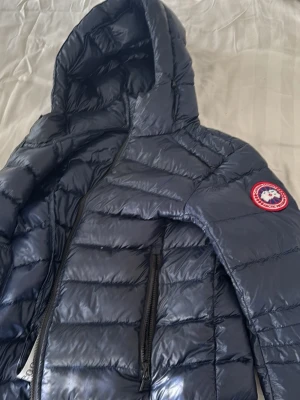 Mörkblå dunjacka från Canada Goose - Snygg mörkblå dunjacka från Canada Goose med huva och dragkedja framtill. Jackan har två sidofickor med dragkedja och den klassiska loggan på ärmen. Perfekt för kalla vinterdagar och riktigt stilren look. Äkta priset kan diskuteras.