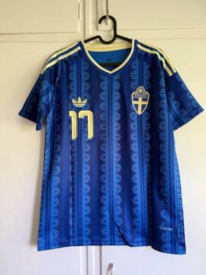 Blå Sverige matchtröja Adidas - Säljer en blå Sverige matchtröja från Adidas med gula detaljer och nummer 17. Tröjan har GYÖKERES tryckt på ryggen, klassiskt mönster och tre gula ränder på axlarna. Tillverkad i lätt och ventilerande material, perfekt för fotboll eller träning.