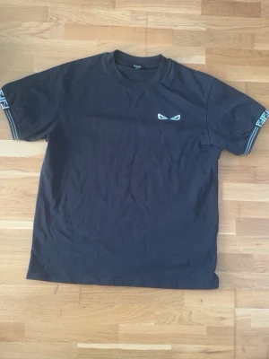 Svart Fendi t-shirt med vita detaljer - Snygg svart t-shirt från Fendi med vita logodetaljer på ärmarna och ett litet ögonmotiv på bröstet. Klassisk passform och rund halsringning. Perfekt för dig som gillar streetwear och exklusiva märken.