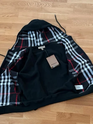 Svart Burberry hoodie med rutigt foder - Säljer en svart hoodie från Burberry med klassiskt rutigt foder i huvan och insidan. Hoodien har dragsko i huvan och är i mjukt material, perfekt för en clean och stilren look. Ikonisk Burberry-tag kvar. Passar dig som gillar exklusiva streetwear vibes.