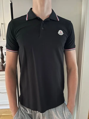 Svart pikétröja från Moncler - Snygg svart pikétröja från Moncler med klassisk krage och knappslå. Tröjan har vita och röda detaljer på krage och ärmslut samt Moncler-logga på bröstet. Perfekt till jeans eller shorts för en clean look. Storlek L men sitter bäst på M