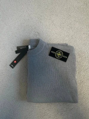 Grå stickad tröja från Stone Island - Snygg grå stickad tröja från Stone Island med klassisk rund hals och ribbade muddar. Tröjan har den ikoniska Stone Island-loggan på vänster ärm och är tillverkad i mjukt material som känns skönt mot huden. Perfekt för dig som gillar stilrena plagg med streetkänsla. Helt ny och aldrig använd. Fraktar samma dag eller nästa📦🚚 Aktiv hela tiden✅ hör av dig om du undrar något eller vill ha fler bilder⁉️