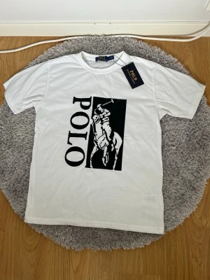 Vit t-shirt från Polo Ralph Lauren - Snygg vit t-shirt med stort svart tryck framtill som visar en polospelare och texten POLO. Klassisk rund halsringning och korta ärmar. Tillverkad i mjuk bomull för en skön känsla och stilren look.