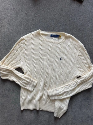 Vit kabelstickad tröja Polo Ralph Lauren - Kabelstickad vit tröja från Polo Ralph Lauren med rund halsringning och broderad marinblå logga på bröstet. Tröjan har långa ärmar och ribbade muddar vid ärmslut och nederkant. Klassisk och tidlös design i mjukt material. Daily wear.(pris kan diskuteras) (är stor i storlek)