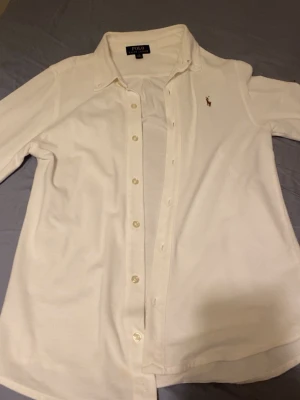 Vit skjorta från Polo Ralph Lauren - Klassisk vit skjorta från Polo Ralph Lauren med knappar framtill och broderad logga på bröstet. Skjortan har lång ärm och en tidlös design som passar till många olika stilar. Tillverkad i mjukt bomullstyg för en bekväm känsla. Storlek passar 14-15 år 