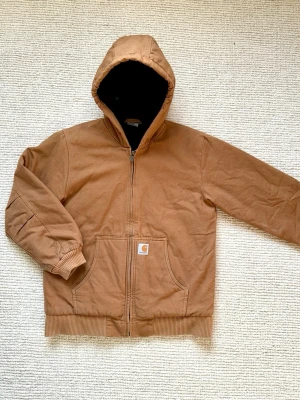 Carhartt Active Jacket – Strl L (passar mindre) - Säljer en snygg Carhartt active jacket i klassisk brun färg med svart innerfoder. Märkt storlek L men passar mer som S/M enligt mig. (SE BILDER FÖR MÅTTEN)  Väldigt clean modell som funkar perfekt till streetwear/workwear fits.  * Märke: Carhartt * Färg: Brun/Camel * Storlek: L (passar mindre) * Skick: Bra skick * Fodrad med huva * Kraftig kvalitet och varm jacka  Säljes eftersom den är för liten för mig.  Skicka gärna meddelande vid frågor eller fler bilder.  