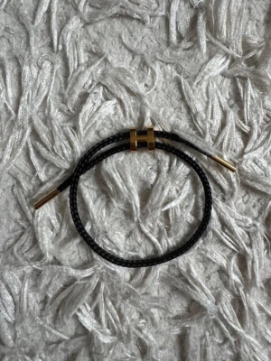 Svart armband med gulddetaljer - Snyggt svart armband i flätat läder med guldfärgad H-formad detalj och guldfärgade ändar. Stilren och modern design som passar perfekt till din vardagsstil. Justerbar storlek tack vare låset i mitten.